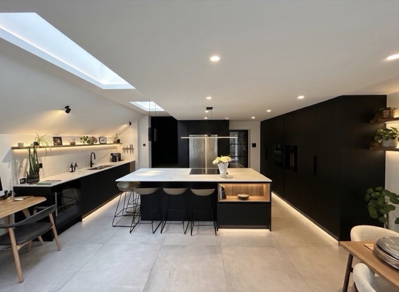 Open-plan kitchen with matt black handleless cabinetry, a white marble-topped island with three stools, an integrated downdraft extractor and two skylights overhead.