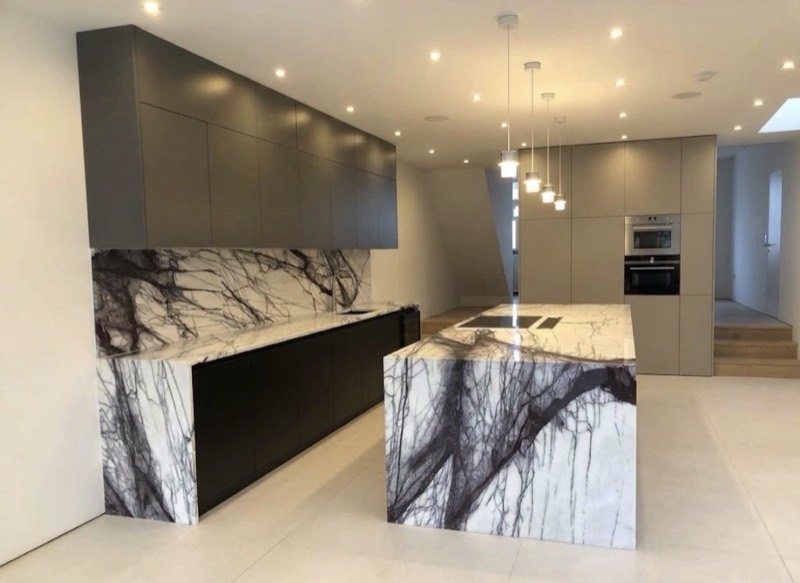 Handleless taupe kitchen with a heavily veined white and purple marble island and matching splashback, beside a run of integrated ovens.
