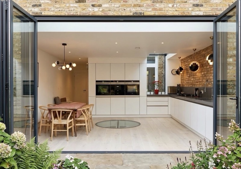 White handleless kitchen in a rear extension, seen through open bifold doors from the garden, with exposed brick, pendant lights and a dining table.
