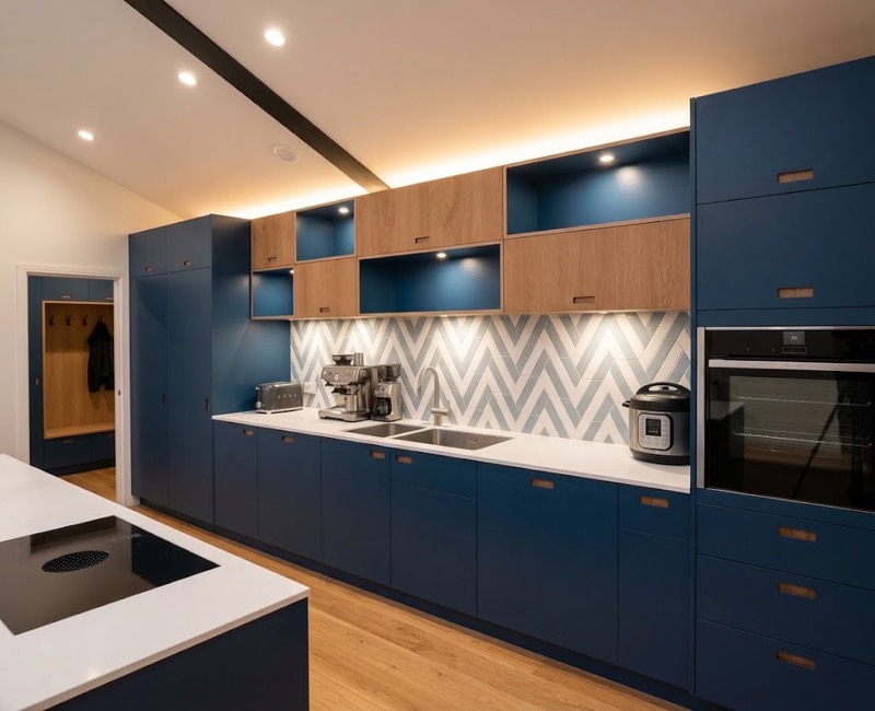 Navy blue kitchen with oak open shelving, a grey chevron-tiled splashback, white worktops and an integrated oven, with a boot room visible beyond.