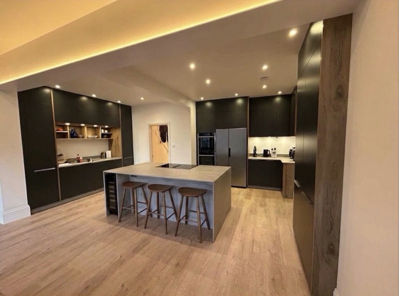 Dark green handleless kitchen with oak open shelving, a concrete-topped island with three wooden stools, and an American fridge freezer.